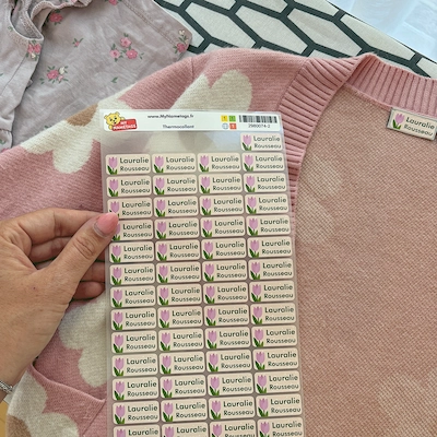 Mão segurando uma cartela de etiquetas termocolantes em frente a um casaco de tricô rosa etiquetado